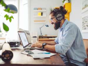 Man wearing headphones and smiling as he types on a laptop in a bright, homey office.