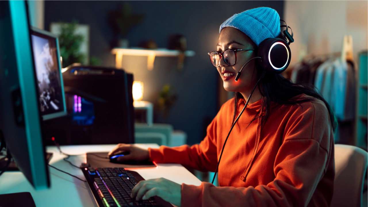 A woman wearing an orange shirt, white beanie with headphones and glasses on uses her computer