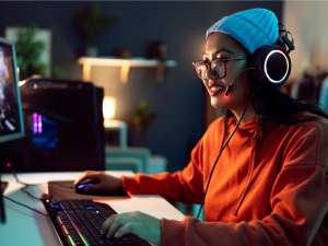 A woman wearing an orange shirt, white beanie with headphones and glasses on uses her computer