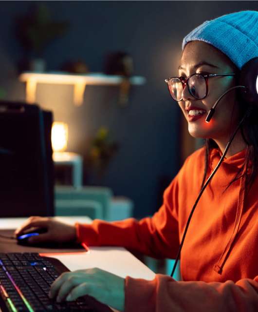 A woman wearing an orange shirt, white beanie with headphones and glasses on uses her computer