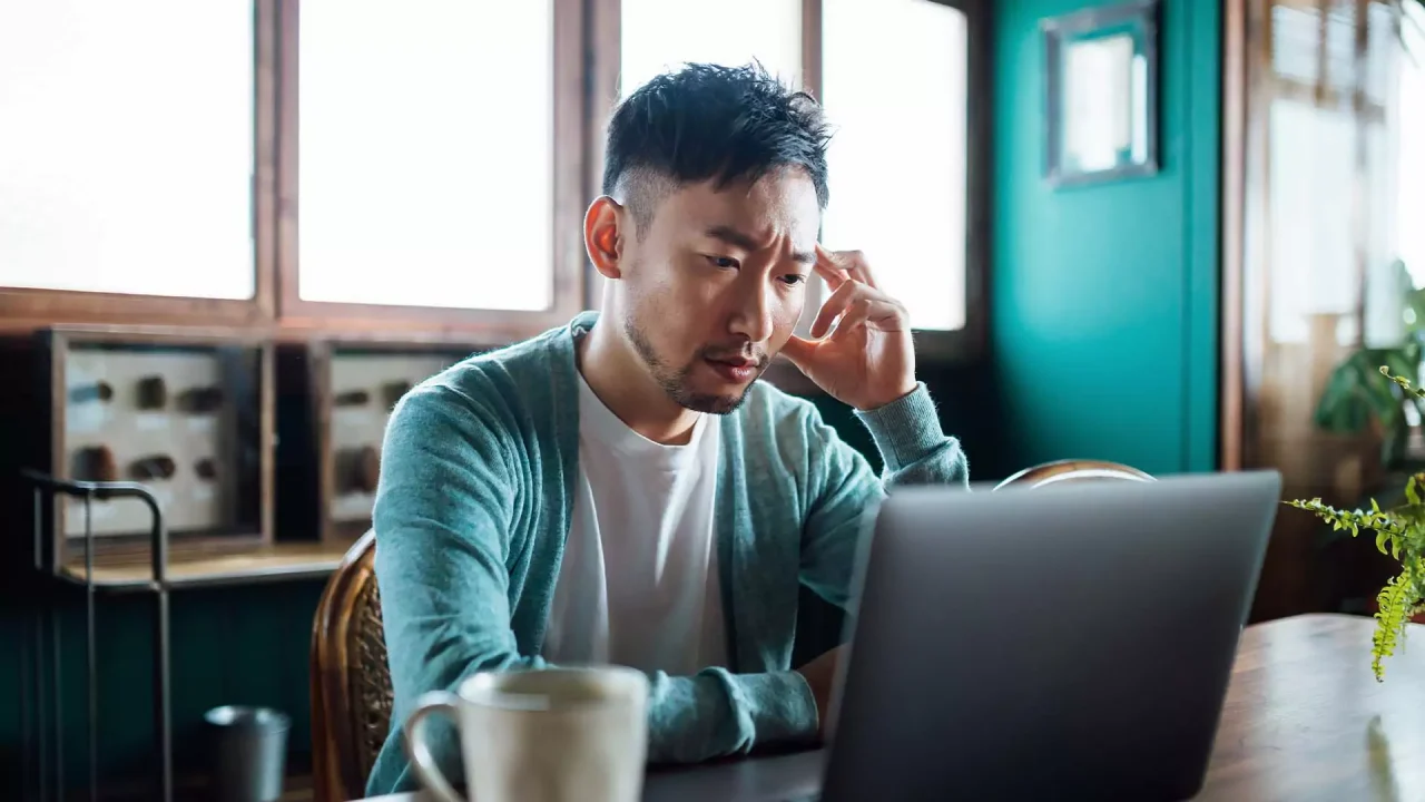 A man sits on his desk looking into his laptop screen and appears stressed.