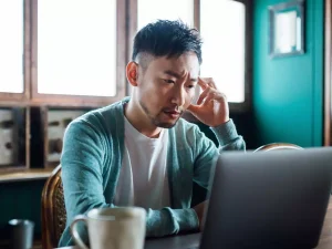 A man sits on his desk looking into his laptop screen and appears stressed.